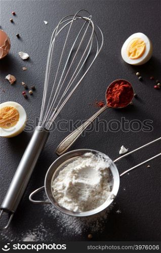 A set of spoon with pepper, sieve with flour, metal whisk and boiled eggs on a black concrete background. Preparation for cooking. Flat lay. Sieve with flour, spoon with red pepper, a metal whisk and halves of a boiled egg on a black concrete background. Concept preparation. Flat lay