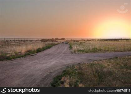 A setting sun fills the sky over the prairie with a dirt road branching in two directions.