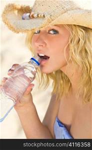 A sexy and beautiful young blond woman sitting at the beach drinking a bottle of water with golden sand behind her