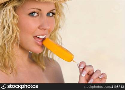 A sexy and beautiful young blond woman sitting on the beach sucking an orange popsicle with golden sand and the sea behind her