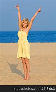 A sexy and beautiful young blond woman stretches up to a blue sky while standing barefoot on a golden beach with a deep blue sea behind her.