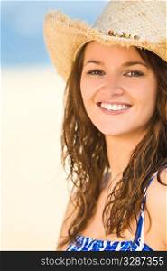 A sexy and beautiful young brunette woman in a cowboy hat, happy and smiling at the beach with golden sand and the sea behind her