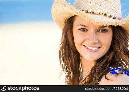 A sexy and beautiful young brunette woman sitting at the beach wearing a straw cowboy hat with golden sand and the sea behind her