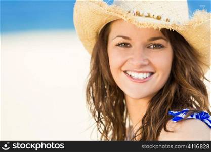 A sexy and beautiful young brunette woman sitting at the beach wearing a straw cowboy hat with golden sand and the sea behind her