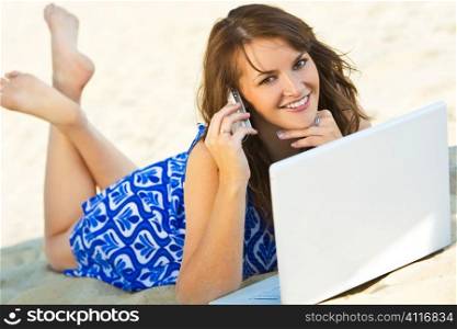 A sexy and beautiful young brunette woman using her laptop and cell phone on the beach