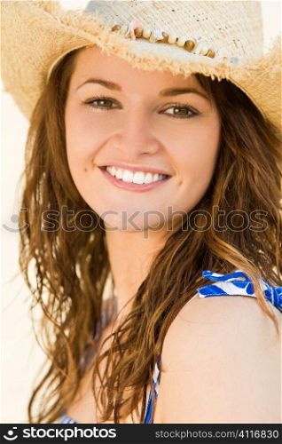 A sexy and beautiful young brunette woman wearing a cowboy hat at the beach with golden sand and the sea behind her