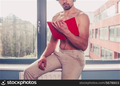 A sexy shirtless young man is sitting on windowsill and reading