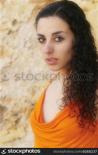 A sexy woman wearing an orange top poses against a stony wall.