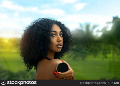 A sexy young black female with long black curly hair   beautiful makeup posing by herself in tropical surroundings while holding a box of powder foundation.