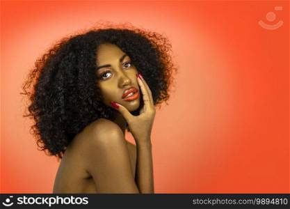 A sexy young black female with long black curly hair, beautiful makeup, popping orange lip stick   perfectly manicured nails posing by herself in front of an orange background.