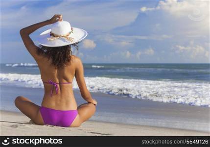 A sexy young brunette woman or girl wearing a bikini and sun hat sitting on a deserted tropical beach with a blue sky