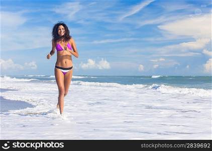 A sexy young brunette woman or girl wearing a bikini running through the surf on a deserted tropical beach with a blue sky