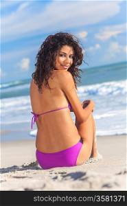 A sexy young brunette woman or girl wearing a purple bikini and ssitting on a deserted tropical beach with a blue sky