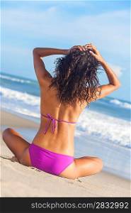 A sexy young brunette woman or girl wearing a purple bikini sitting on a deserted tropical beach with a blue sky