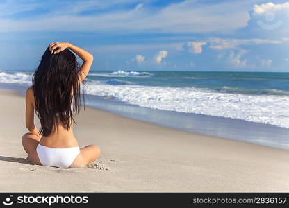 A sexy young brunette woman or girl wearing a white bikini sitting on a deserted tropical beach with a blue sky