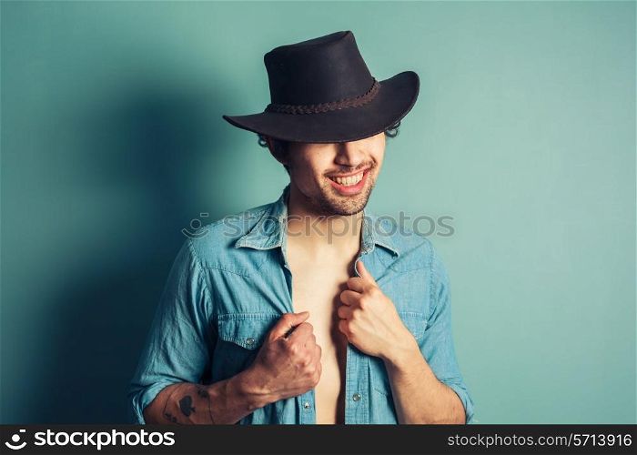 A sexy young cowboy is standing by a blue wall with his shirt unbuttoned