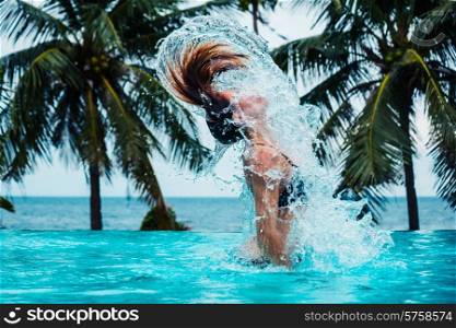 A sexy young woman is doing a hairflip and creating splashes in a swimming pool with palm trees and the ocean in the background