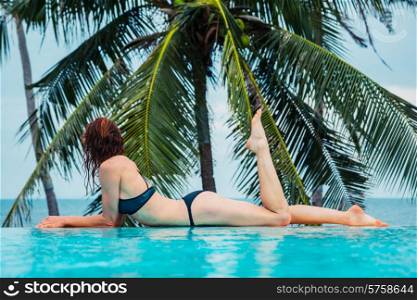 A sexy young woman is relaxing by a swimming pool with palm trees and the ocean in the background