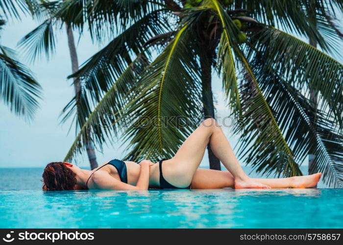 A sexy young woman is relaxing by a swimming pool with palm trees and the ocean in the background