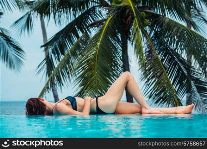 A sexy young woman is relaxing by a swimming pool with palm trees and the ocean in the background