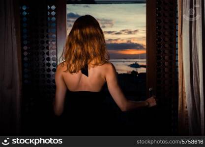 A sexy young woman wearing a dress is looking out at the sunset through her french doors