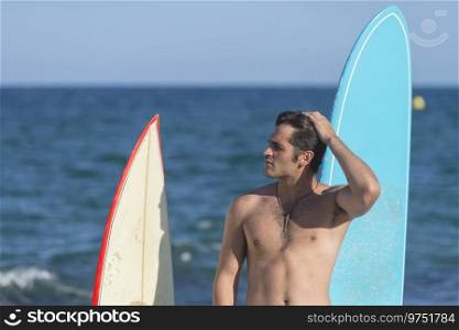 A shirtless man surfer standing on the beach holding a surfboard - looking down