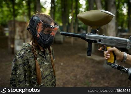 A shot from a paintball gun in the girl&rsquo;s face in mask, playground in the forest on background. Extreme sport with pneumatic weapon and paint bullets or markers, military team game outdoors. Shot from paintball gun in the girl&rsquo;s face in mask
