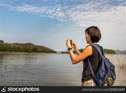 A side view of a young asia woman using her smartphone up and pointing it to take pictures view of lake shore with mountains range in background. Technology and holiday travel, lifestyle, Nature, No focus, specifically.