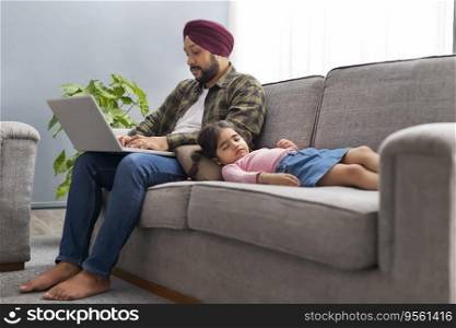 A SIKH MAN WORKING ON LAPTOP WHILE DAUGHTER COMFORTABLY SLEEPING NEAR HIM ON SOFA