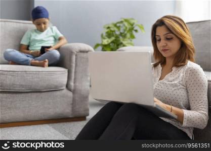 A SIKH SON SITTING ON SOFA WITH  CROSSED LEGS USING MOBILE PHONE WHILE MOTHER WORKS ON LAPTOP SITTING NEARBY