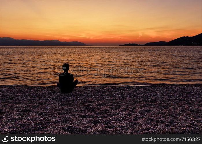 A silhouette of a teenager who sits on a beach of small pebbles of the seashore and watches a beautiful, bright sunset on the Corinthian Gulf, Loutraki, Greece,. Silhouette of a teenager sitting on the seashore and watching a beautiful, bright sunset on the Gulf of Corinth.