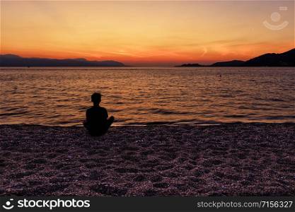A silhouette of a teenager who sits on a beach of small pebbles of the seashore and watches a beautiful, bright sunset on the Corinthian Gulf, Loutraki, Greece,. Silhouette of a teenager sitting on the seashore and watching a beautiful, bright sunset on the Gulf of Corinth.