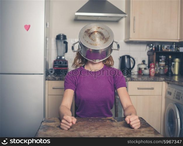 A silly young woman is sitting at a table in a kitchen with a pot on her head