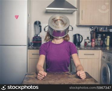 A silly young woman is sitting at a table in a kitchen with a pot on her head