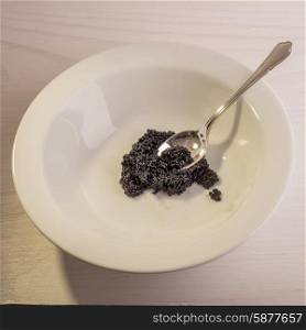 A silver tea spoon lies inside a white ceramic bowl , dipped in to some black caviar, all situated on a white wooden surface.