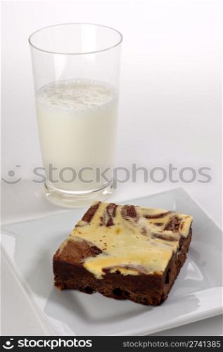A single cream cheese brownie square shot in the studio on a white plate with a glass of milk in the background.