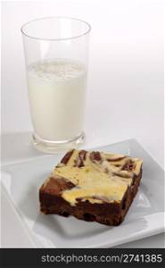 A single cream cheese brownie square shot in the studio on a white plate with a glass of milk in the background.