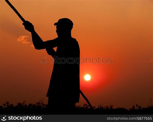 A skilled poler pushes his dugout boat forward in the Okavango Delta, while standing upright in the boat, and seen as a silhouette, arms stretched out, while holding the pole in both hands, behind him the yellow glow of the sun setting in a beautiful red orange sky.