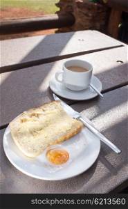 A slice of freshly baked white bread lies on a plate on an outside table, already spread with butter, with sam marmalade next to it, and a cup of coffee behind.