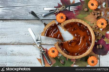 A slice of pumpkin pie with autumn foliage and small pumpkins on rustic white wood. Thanksgiving holiday concept in flat lay format.