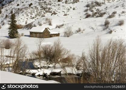A small barn in a rural area near Aspen, Colorado is completely covered with snow. In the foreground, a small stream is running.