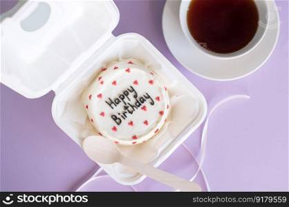 A small bento cake in eco-packaging with a wooden spoon and ribbon next to a cup of tea on a purple background. A small bento cake in eco-packaging with a wooden spoon next to a cup of tea on a purple background