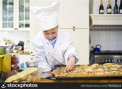A small boy, dressed up as a chef, garnishing the sweet bread he baked in a cluttered kitchen
