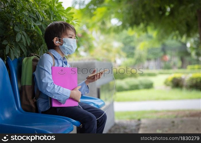 A small boy wearing face mask with a schoolbag sitting alone on the chair under the building of school during covid pandemic.He is using smart phone listening music during waiting.