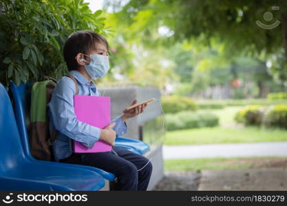 A small boy wearing face mask with a schoolbag sitting alone on the chair under the building of school during covid pandemic.He is using smart phone listening music during waiting.