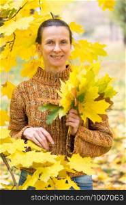 A smiling adult caucasian woman is picking yellow maple leaves in autumn in the park