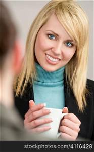 A smiling businesswoman drinking from a mug in a meeting with her colleague out of focus in the foreground