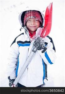 A smiling caucasian child looking at camera, wearing white winter clothing and holding a red snow shovel. Winter season.