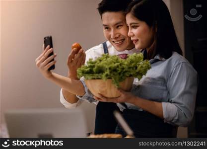 a Smiling Couple of Male and Female Using Phone in the Kitchen. Taking Selfie or Live Streaming via Smartphone while Preparing a Vegan Food. Modern Healthy Lifestyle Concept. Cooking At Home