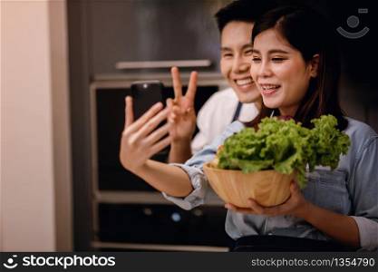 a Smiling Couple of Male and Female Using Phone in the Kitchen. Taking Selfie or Live Streaming via Smartphone while Preparing a Vegan Food. Modern Healthy Lifestyle Concept. Cooking At Home Together